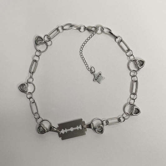 Image shows necklace against white background. The necklace is a steel razor blade connected by handmade chain with heart spacer beads hanging from the large jump rings. The necklace has a 2” extender chain with a star shaped charm tag at the end of it with the caution toxic logo engraved on it 