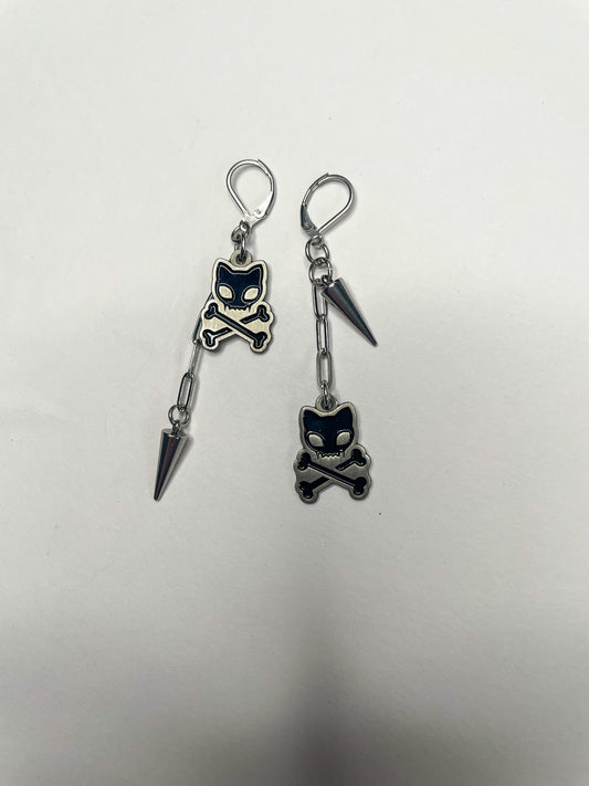 Image shows two earrings against a white background. One earring has a 1” chain ending ina. Silver spike with a shorter connector holding a small .75” black enamel charm depicting a kitty skull and vorssvones. The other earring is the opposite with the longer chain ending in the enamel charm and the shorter connector holding the spike 