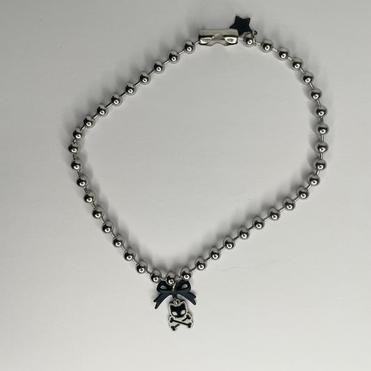 Image shows necklace on a white background. Necklace is a chunky 8mm ball chain— the pendant is a small black matte metal bow connector with a small black enamel cat skull and crossbones charm. The charm measures .85” and near the connector there is a star shaped charm tag with the caution to I. Logo or rawr engraved on it 