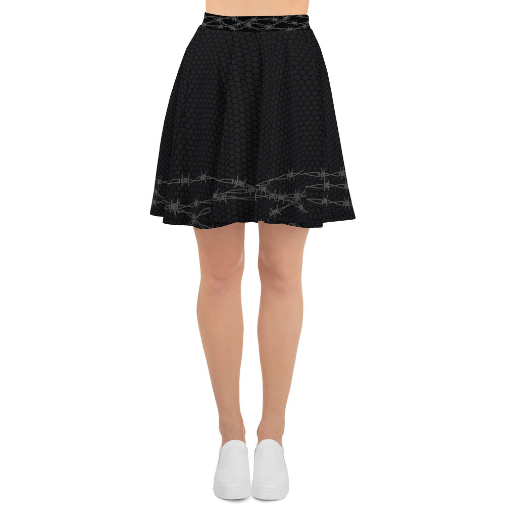 Image shows lower half of female body wearing a skater skirt that ends just above the knee. The skirt is a soft black with chain link fence pattern, with the bottom half of the skirt wrapped in barbed wire design 